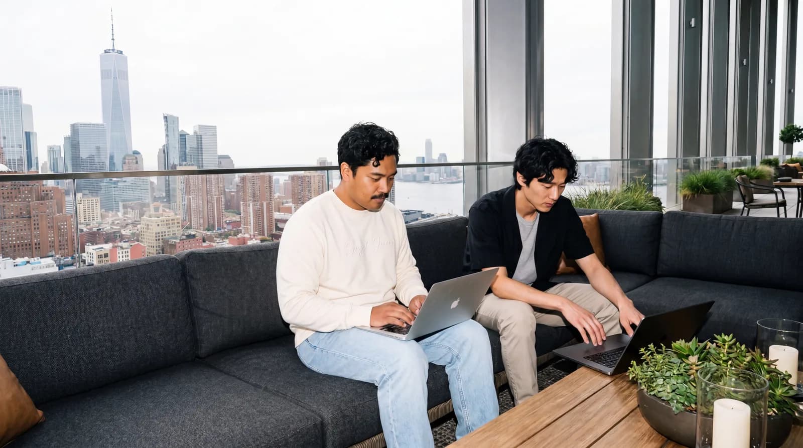 Two people collaborate on a sofa beside skyline windows.