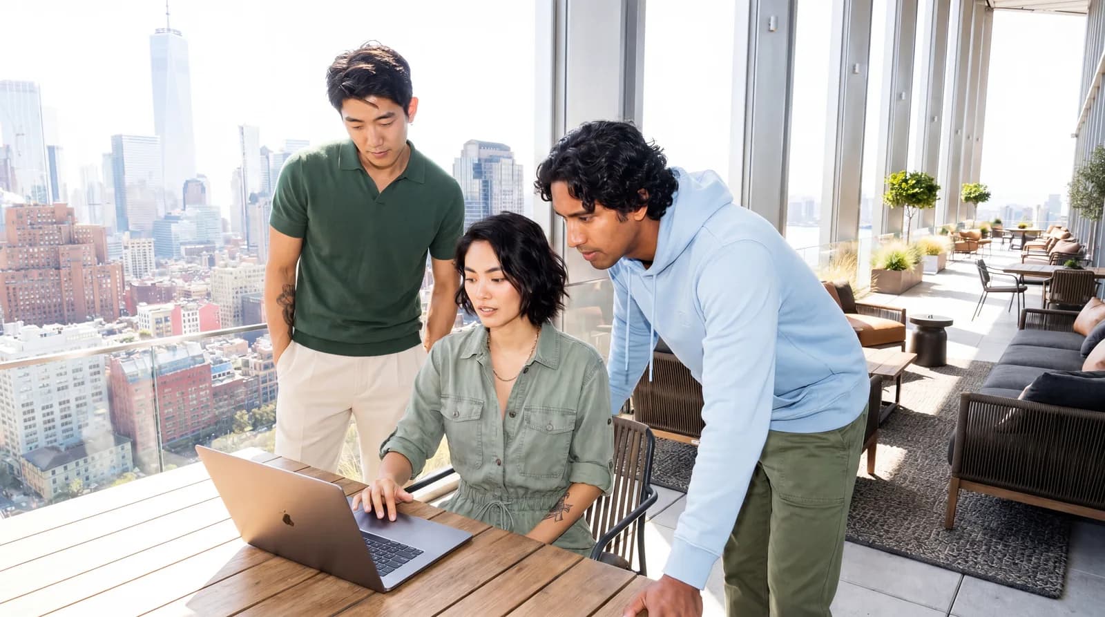 A team collaborates around a laptop beside floor-to-ceiling windows.