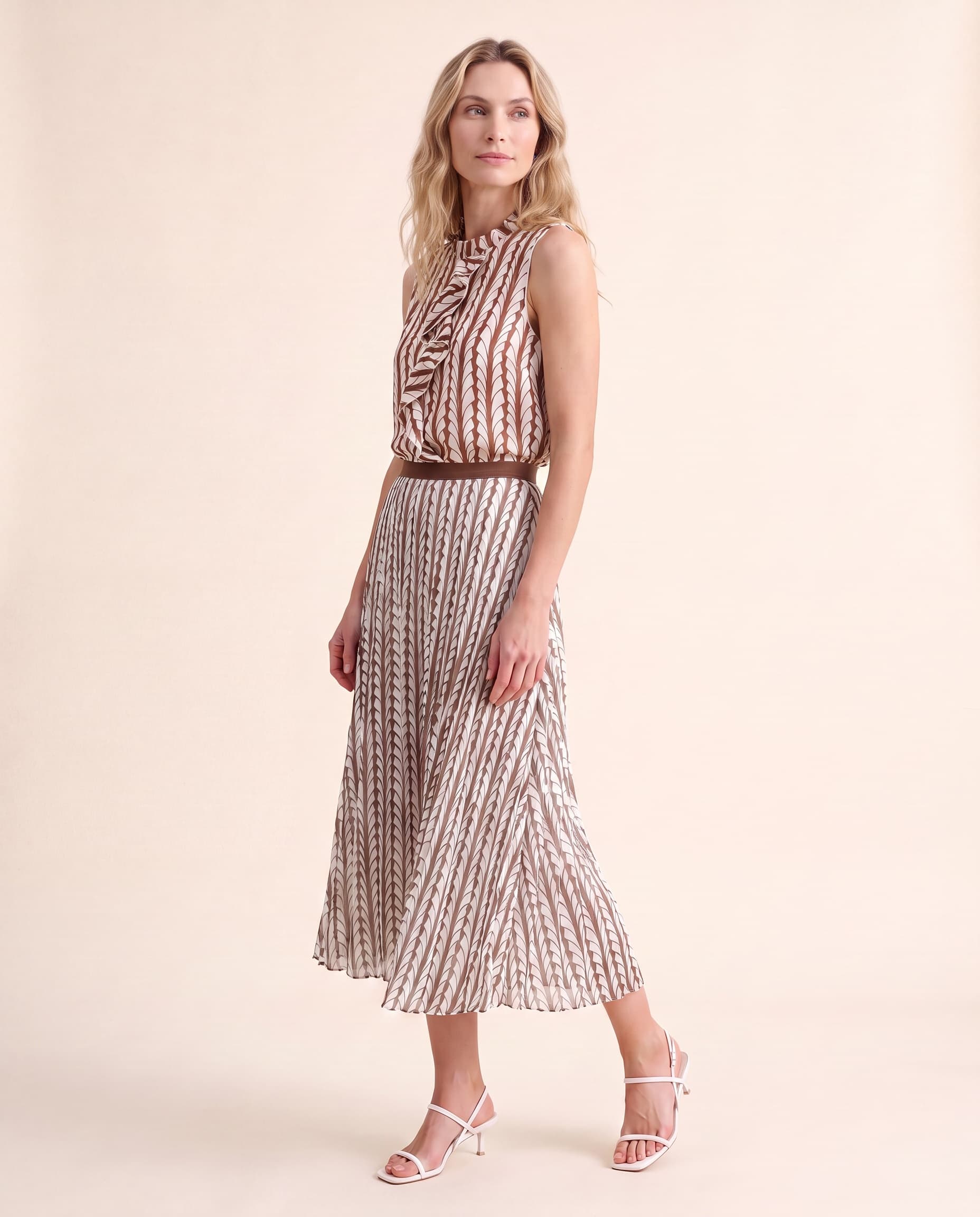 An AI model smiles over her shoulder in the Cartolina Cassie ruffle top and Claudia pleated skirt, both in the thrush print, on a cream studio backdrop.