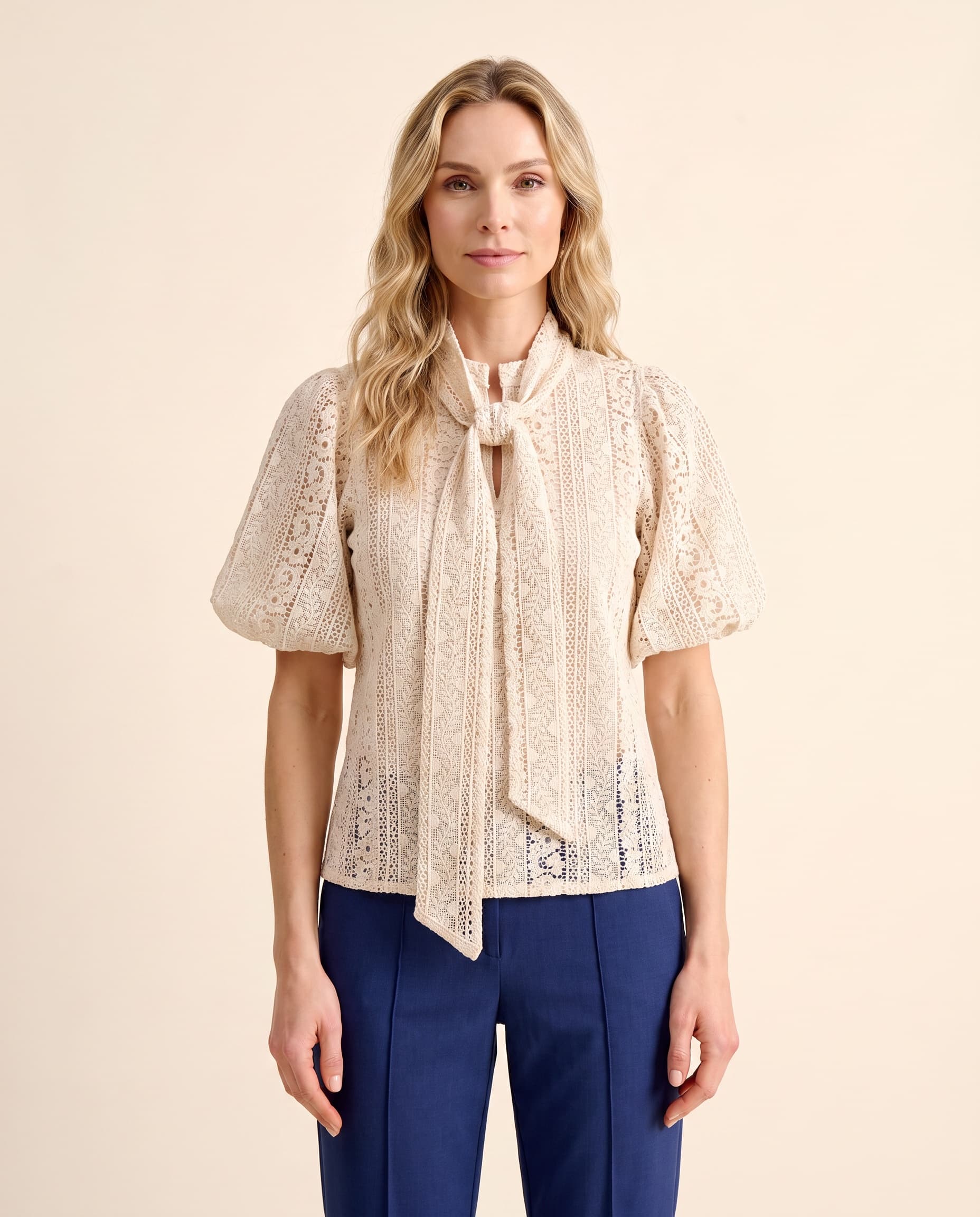An AI model wears the Cartolina Perry tie-neck top in ecru lace with the Beverly pant in navy, standing full length on a cream studio backdrop.