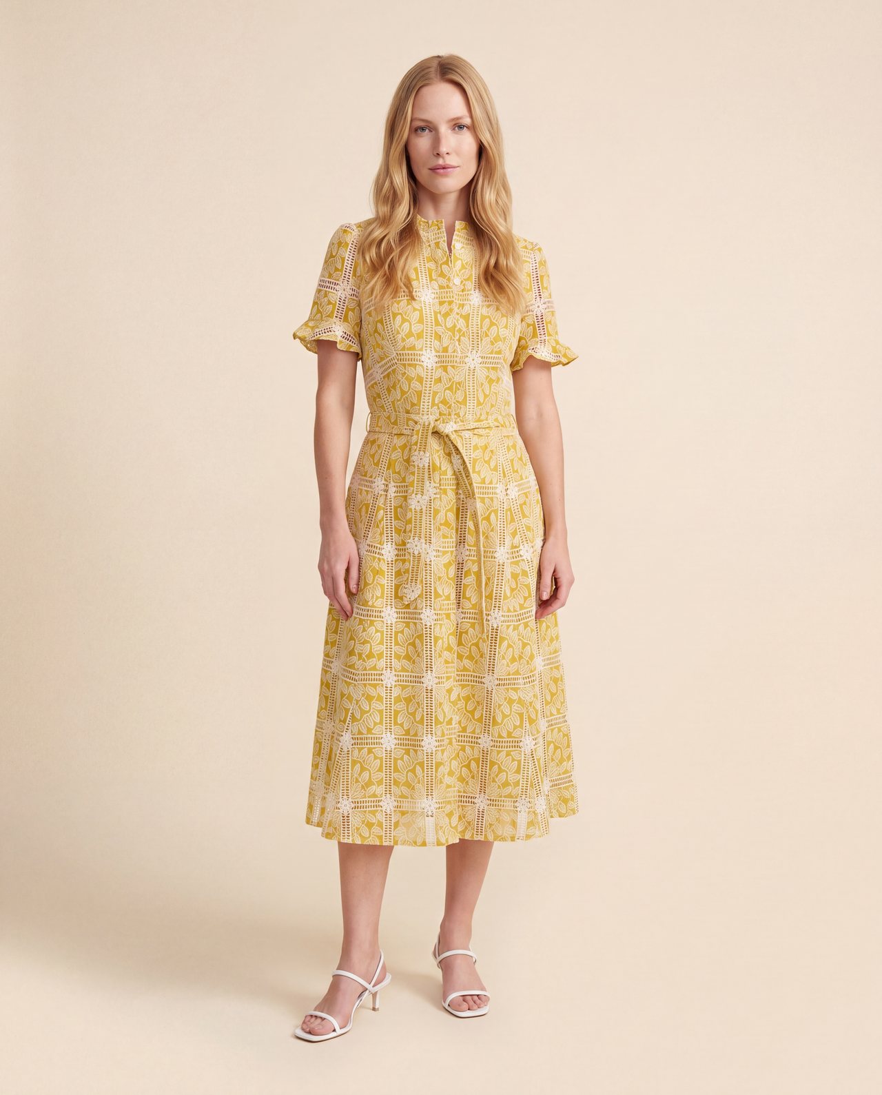 An AI model wears the Cartolina Aria dress in marigold eyelet, tied at the waist with its matching belt, standing on a cream studio backdrop.