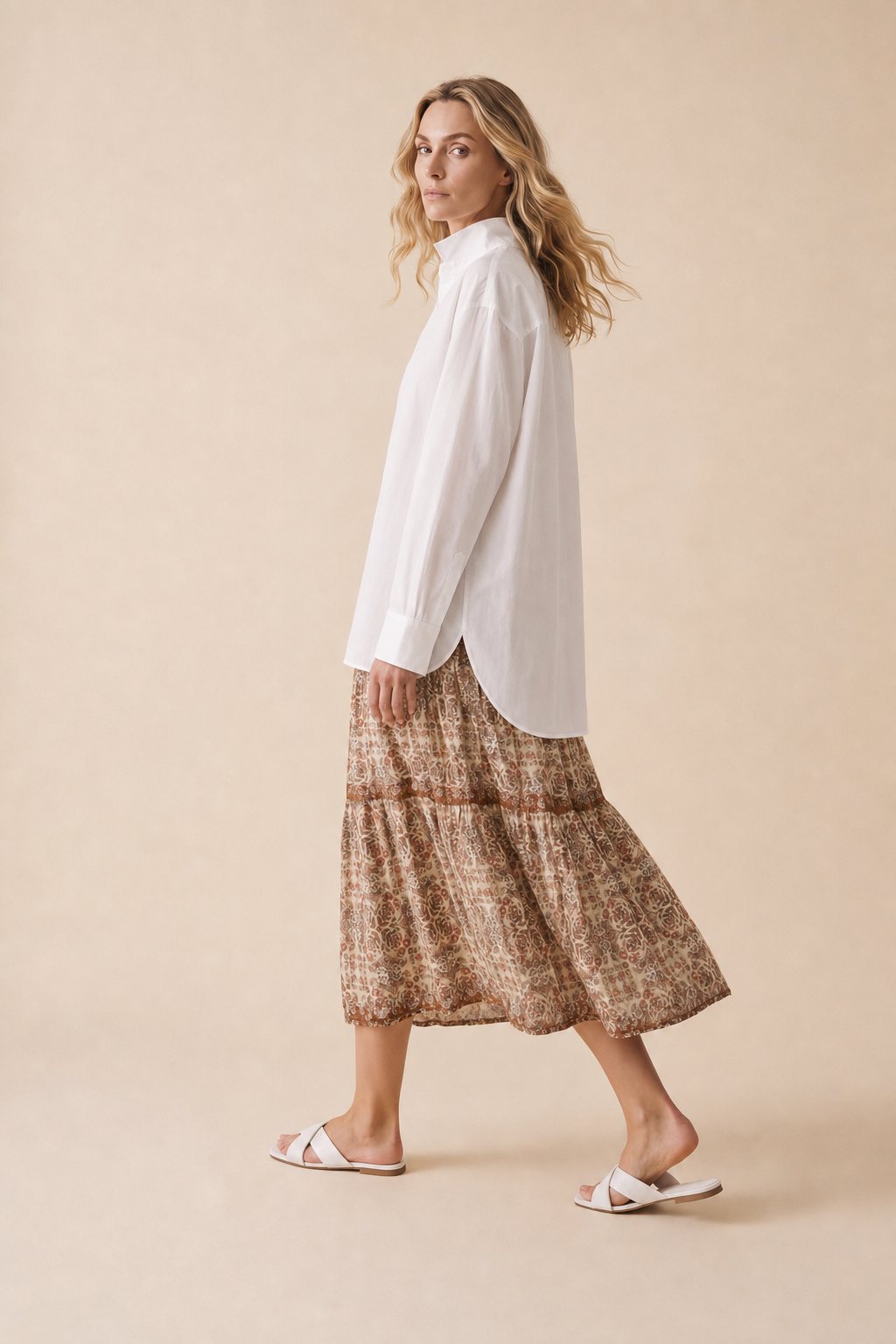 An AI model walks in the Cartolina Betsey shirt in white, worn untucked with its long curved back tail over the Kylie tiered skirt in the boho print.