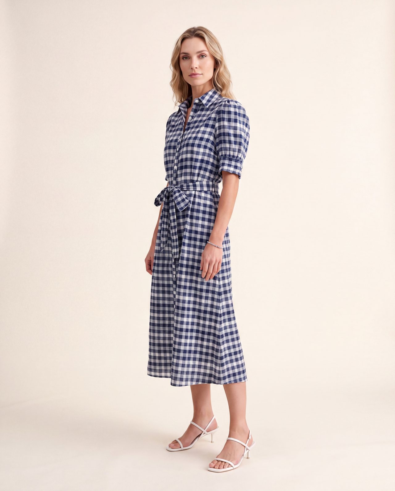 An AI model wears the Cartolina Francesca shirt dress in navy gingham with puff sleeves and a tie belt, standing on a cream studio backdrop.