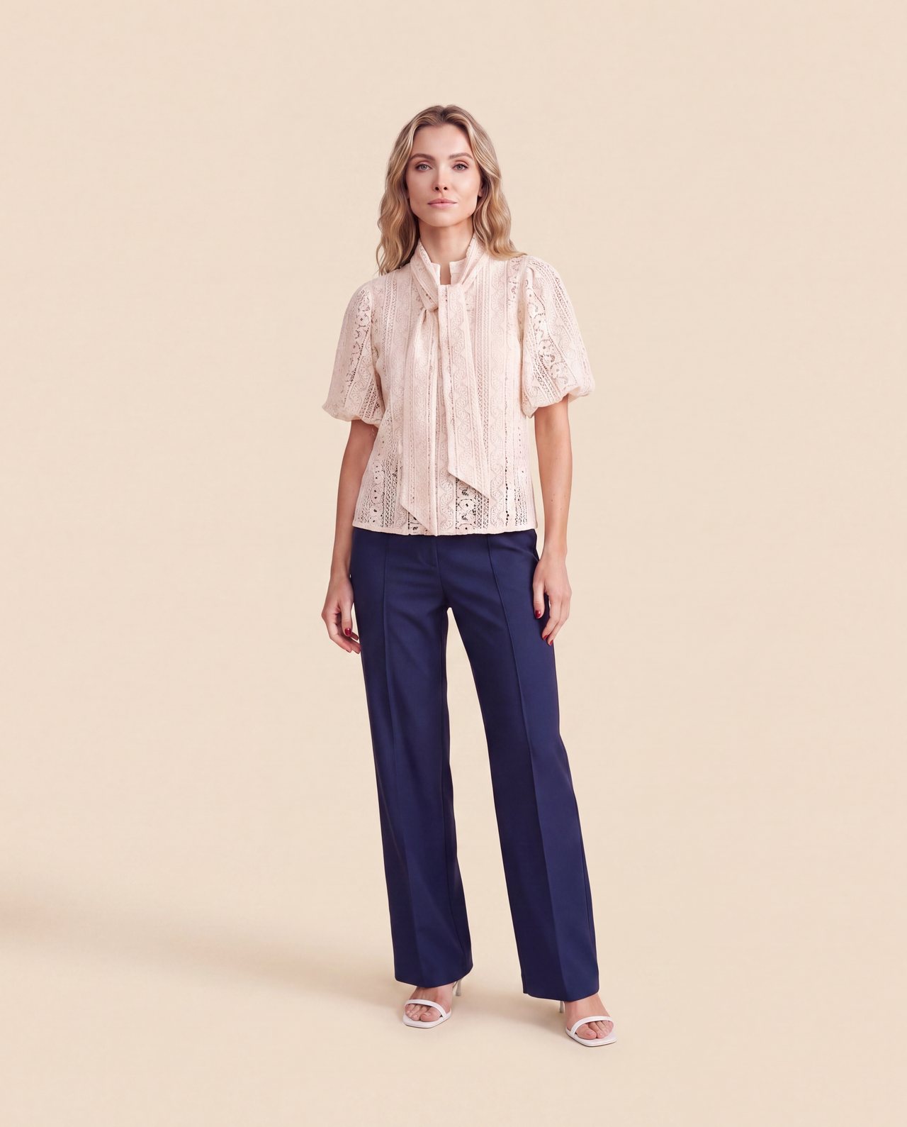An AI model wears the Cartolina Perry tie-neck top in ecru lace with the Beverly pant in navy, standing full length on a cream studio backdrop.