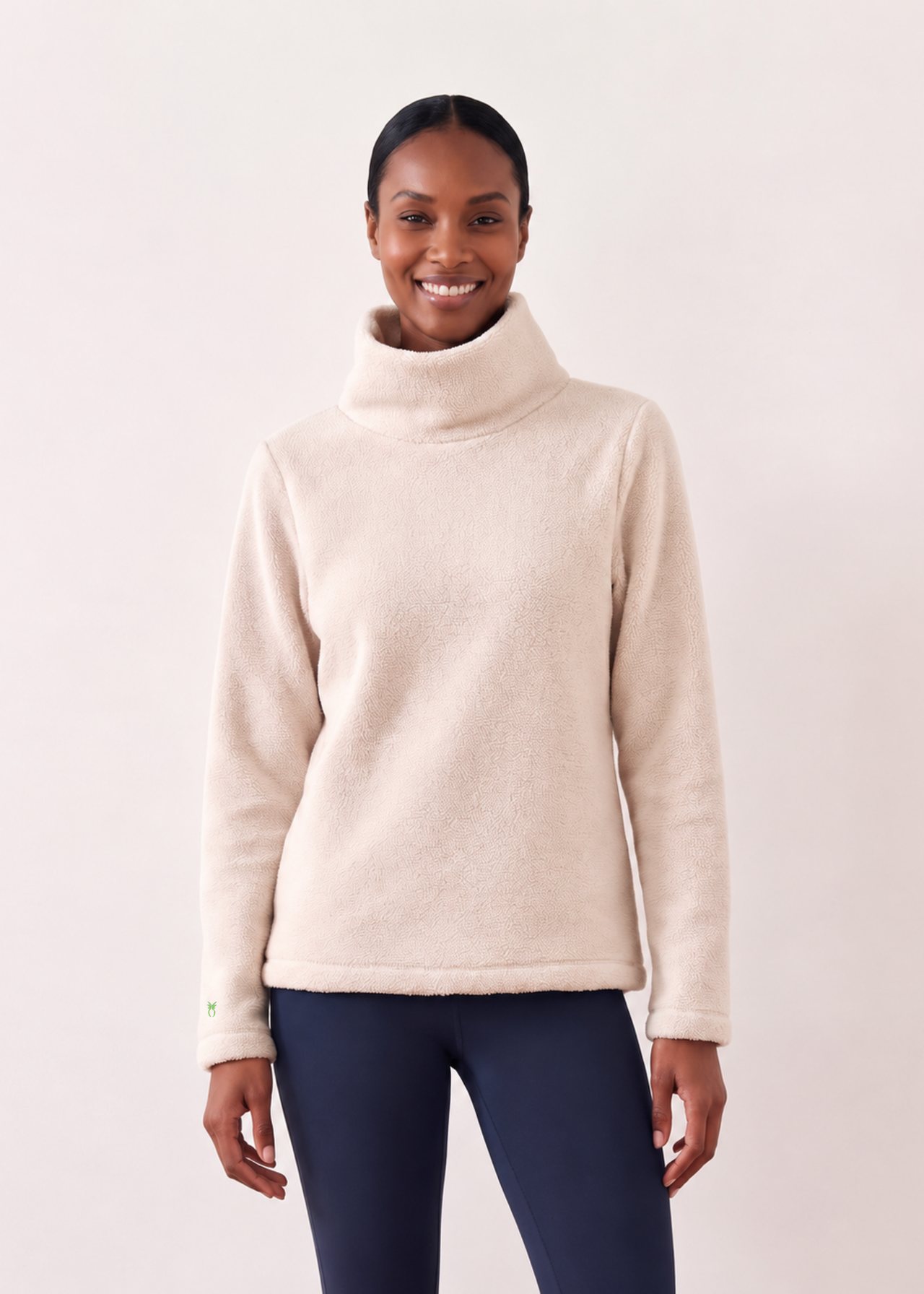 An AI model smiles in the Dudley Stephens Greenpoint turtleneck in oatmeal fleece with navy leggings, against a white studio wall.