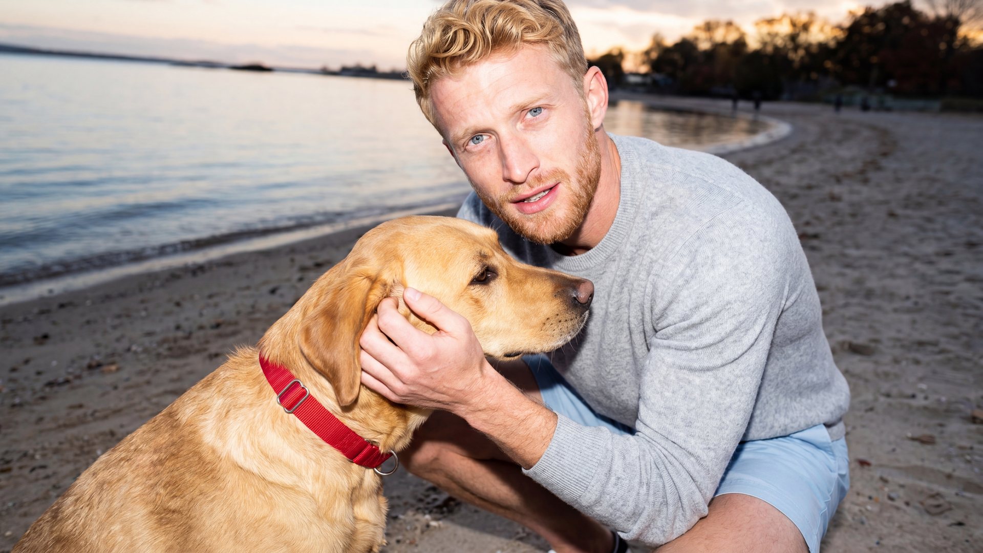 A man in a grey sweater crouches on a sandy beach at sunset and cradles the head of his yellow Labrador.