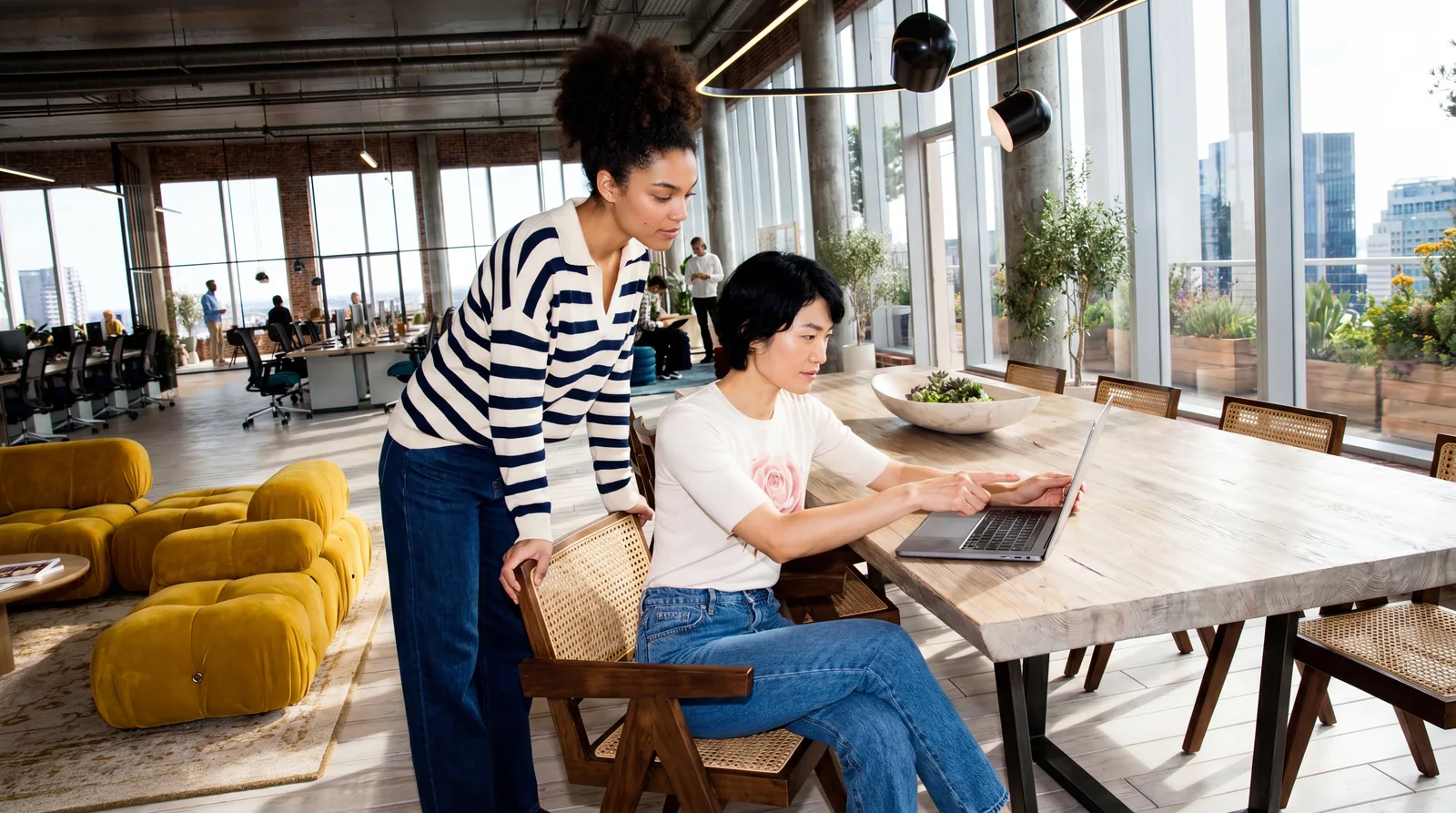 Two people review a laptop at a shared table in a bright workspace.
